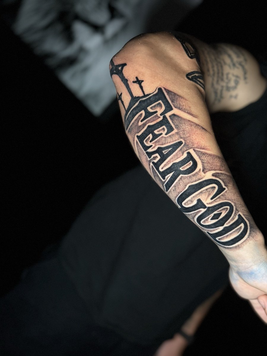 Bold Fear God lettering tattoo across a forearm.